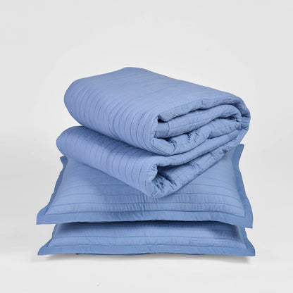 Vliving denim blue quilted cotton bedspread with stripe quilting and border, folded on top of two pillow shams.