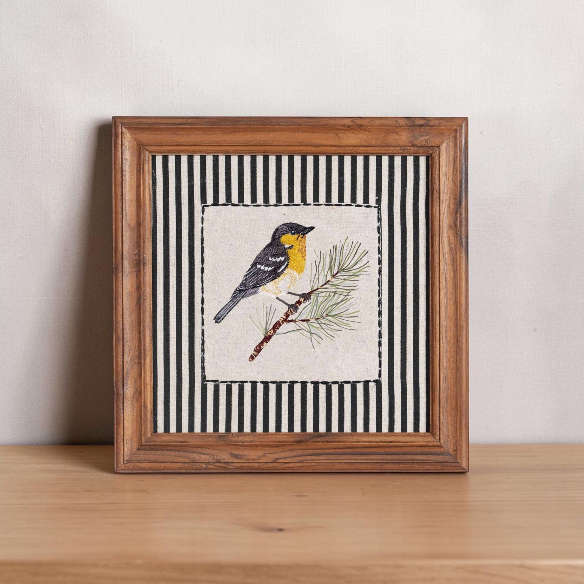 Vliving framed embroidered yellow and grey bird on a pine branch with black and white striped background.