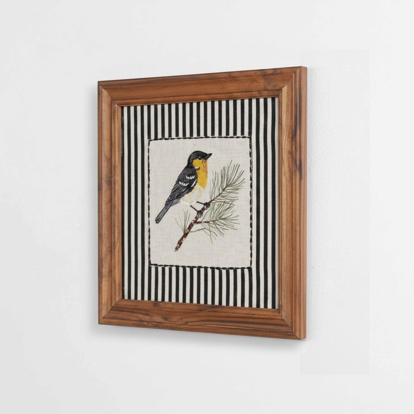 Vliving embroidered yellow and grey bird on a pine branch, framed in wood with black and white striped background.