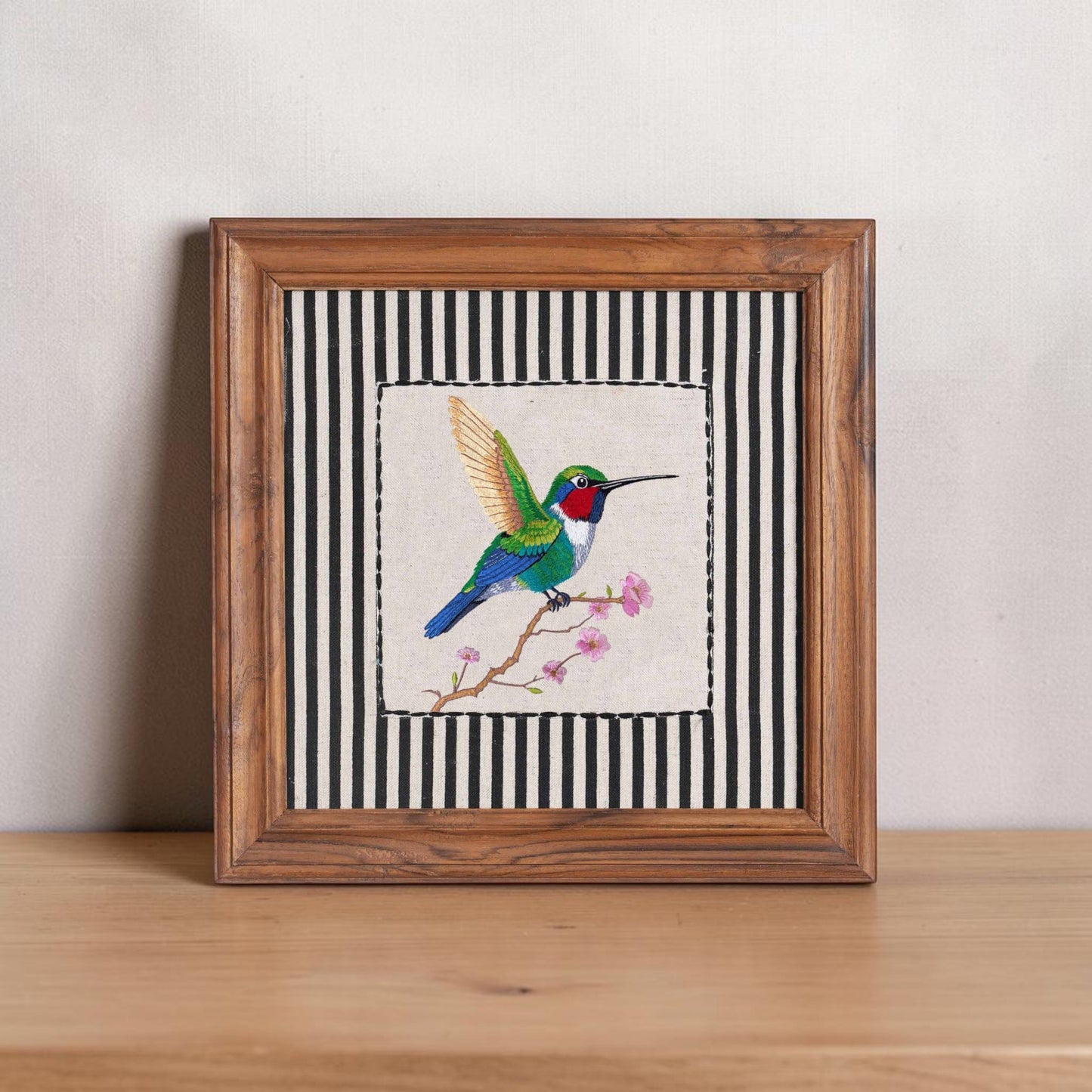 Vliving BOTANICAL hummingbird embroidered wall art with teak wood frame, featuring a colorful bird on a branch with pink blossoms against a black and white striped background.
