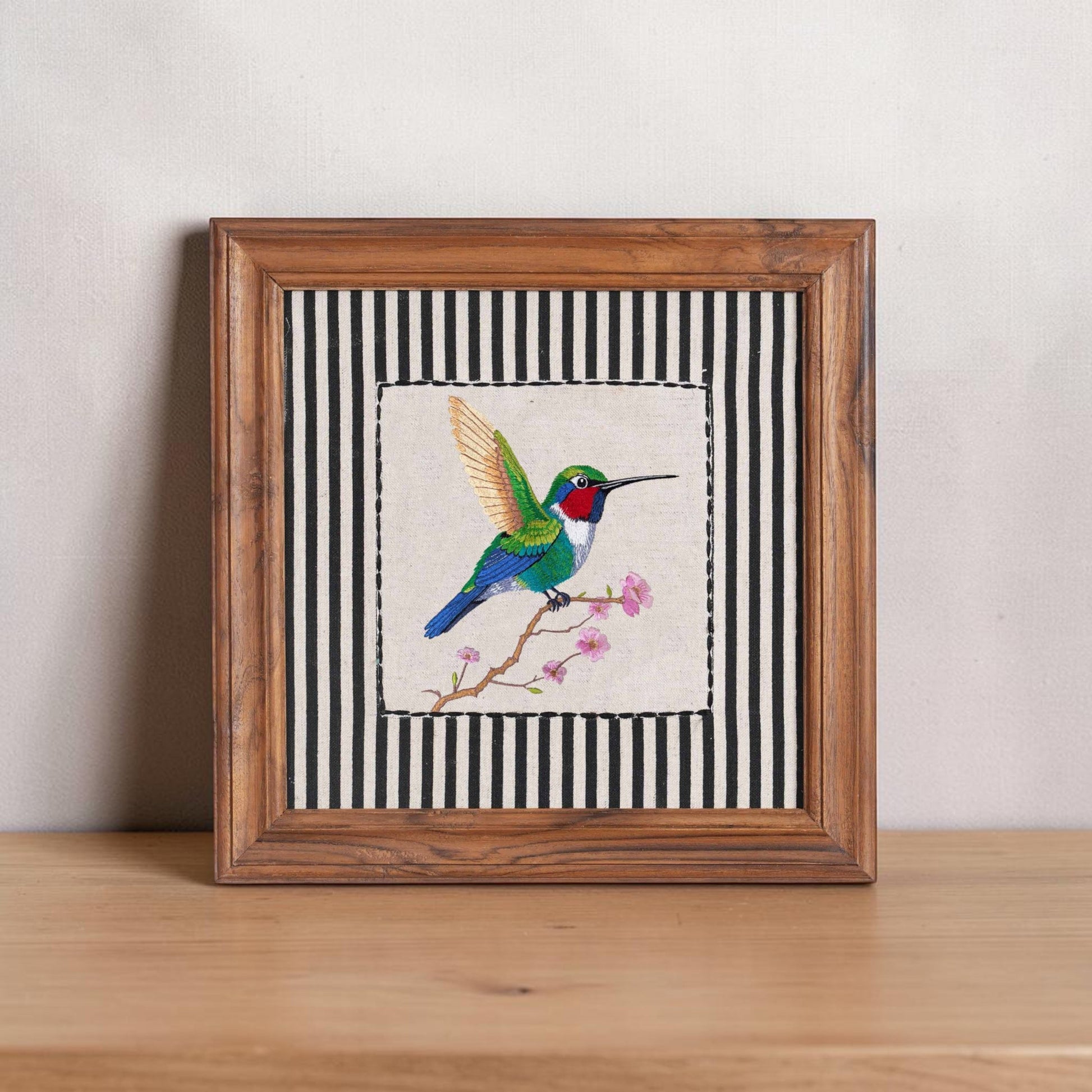 Vliving BOTANICAL hummingbird embroidered wall art with teak wood frame, featuring a colorful bird on a branch with pink blossoms against a black and white striped background.